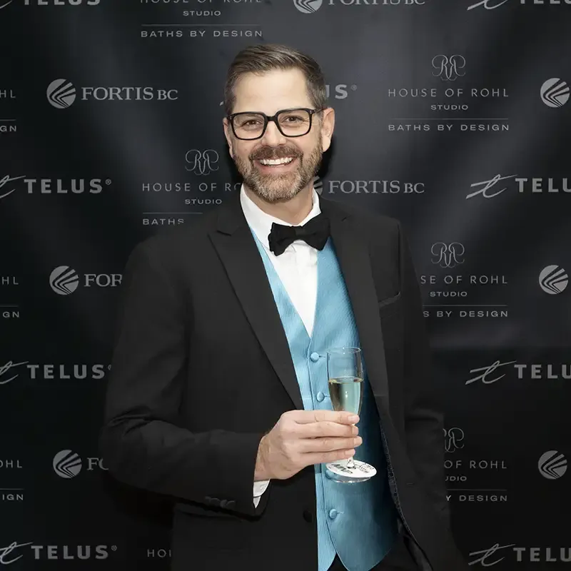 Cliff holding a champagne glass at an event with sponsors backdrop behind